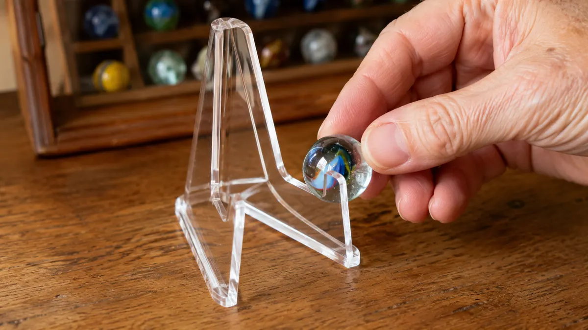 A hand carefully setting a large shooter marble onto the lip of a clear acrylic easel stand