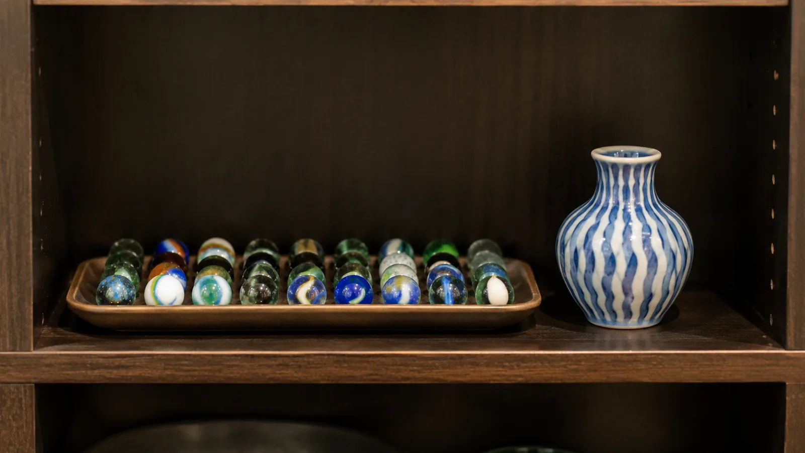 The interior of a lit cabinet shelf showing a row of glass marbles and a small vase spaced evenly apart