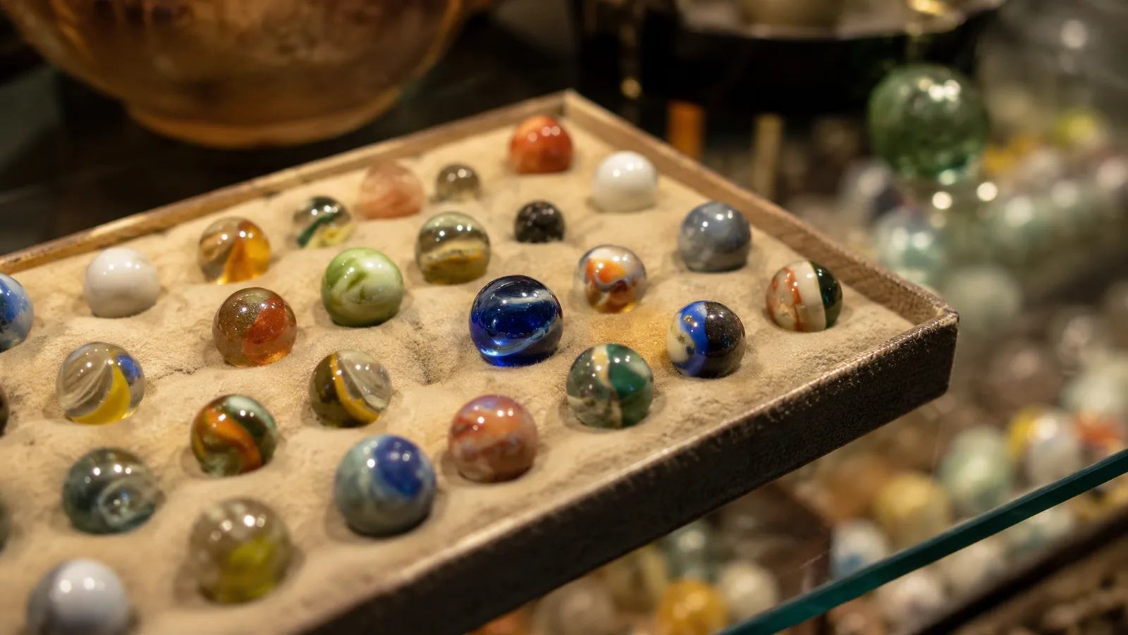 Close view of a padded tray of vintage swirl marbles on a lit glass shelf inside the cabinet, one deep blue swirl marble catching the warm light
