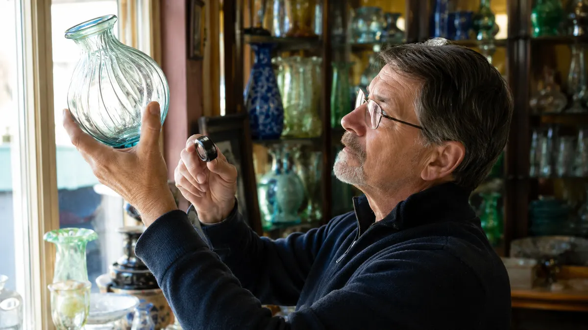 Collector inspecting the seam line of a vintage green glass vase in a small shop near a shelf of glassware