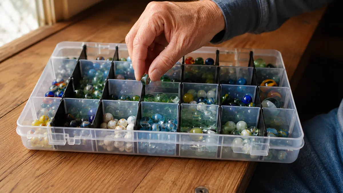 A hand dropping a green cat's eye marble into a filled clear grid organizer tray on a bench